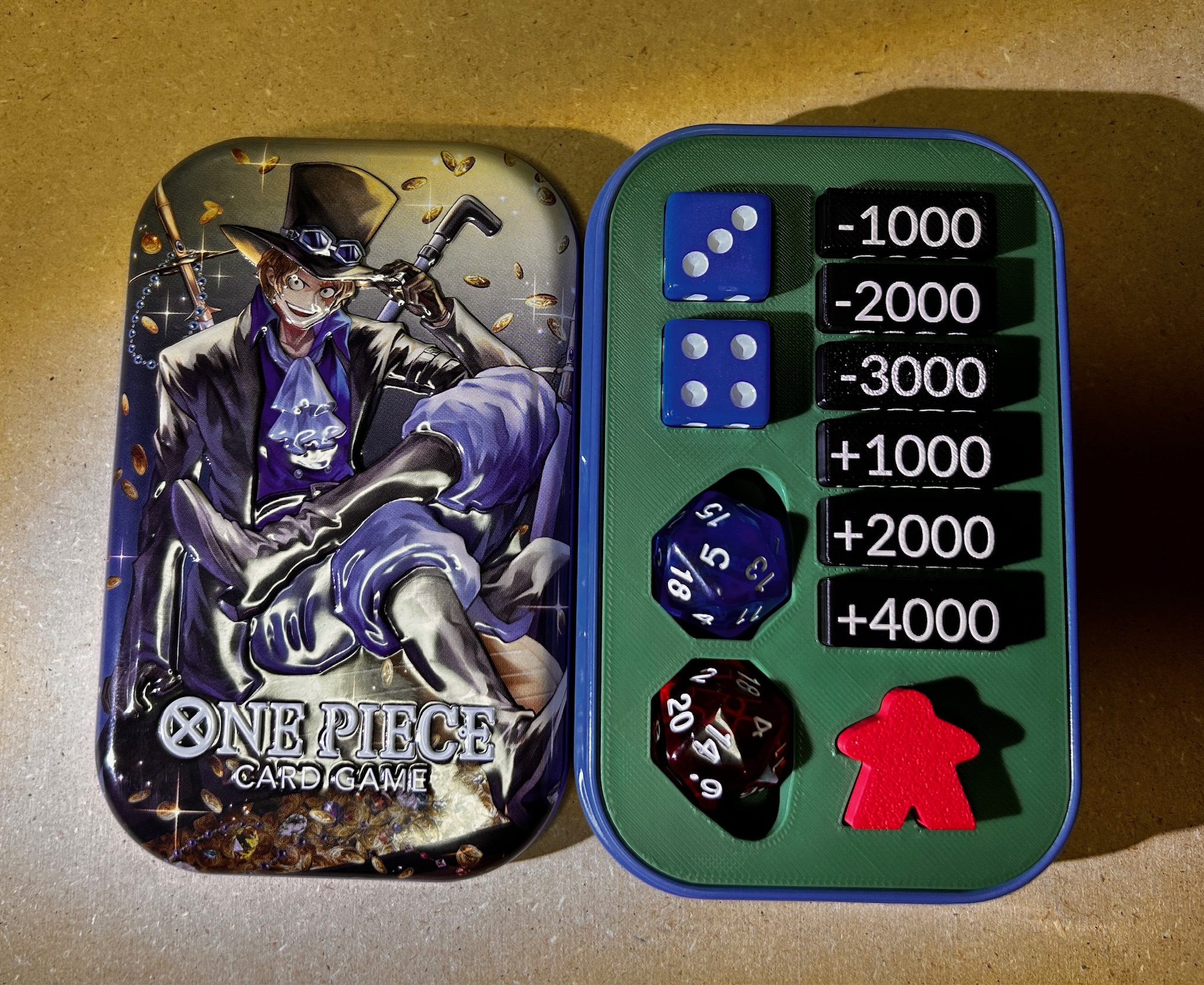 Open One Piece Card Game tin with its illustrated lid beside a fitted green TinForge insert holding game pieces